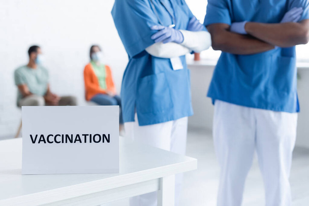 interracial physicians standing with crossed arms near blurred patients in vaccination center - Foto, Imagen
