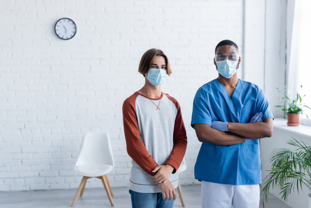 médico afroamericano de pie con los brazos cruzados cerca de hombre joven en máscara médica, concepto de vacunación - Foto, Imagen