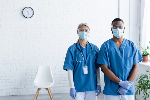 médicos multiculturales en uniforme y máscaras médicas mirando a la cámara en el centro de vacunación - Foto, Imagen