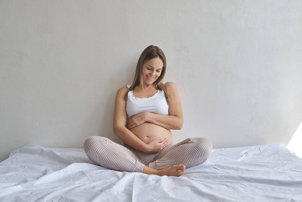 pregnancy, rest, people and expectation concept - happy pregnant woman sitting on bed and touching her belly at home - Fotó, kép