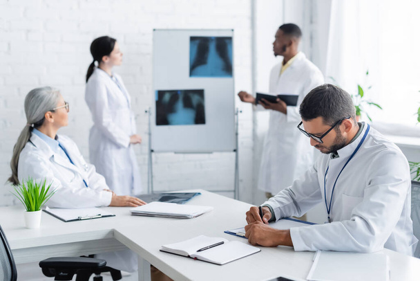 nuori lääkäri kirjoittaa leikepöydälle, kun taas hämärtynyt monikansalliset kollegat puhuvat lähellä keuhkojen röntgenkuvat - Valokuva, kuva