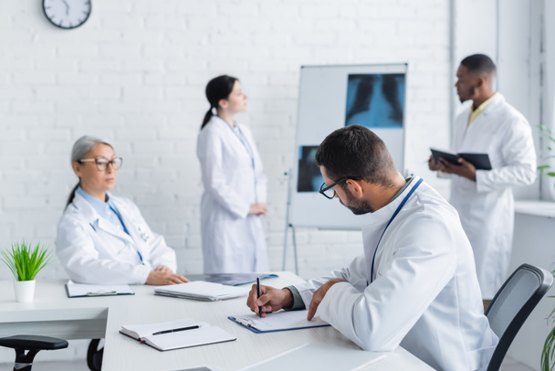joven médico escribiendo en un cuaderno mientras colegas multiculturales hablando cerca de los pulmones rayos X sobre fondo borroso - Foto, imagen