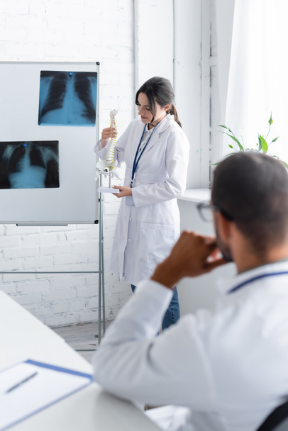 Junge Ärztin hält Wirbelsäulenmodell ordentlich Flipchart mit Röntgenaufnahmen und verschwommenem Kollegen  - Foto, Bild