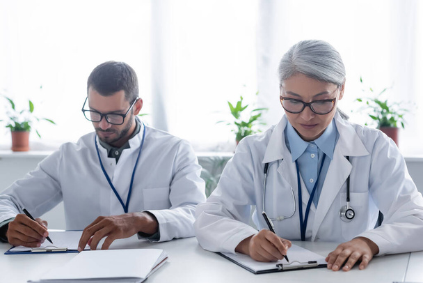 young and mature interracial doctors in eyeglasses writing on clipboards - Foto, immagini
