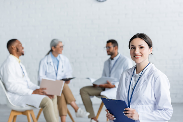 young doctor with clipboard smiling at camera while multiethnic colleagues talking on blurred background - Photo, Image