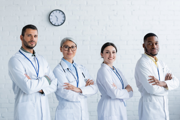 happy multicultural doctors in white coats standing with crossed arms and looking at camera - 写真・画像