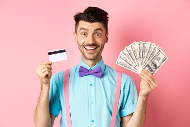 Fröhlicher Kaukasier mit Schnurrbart und Fliege zeigt Plastikkreditkarte mit Geld in Dollar, lächelt in die Kamera, steht über rosa Hintergrund - Foto, Bild