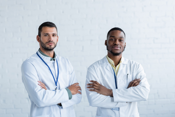 νέοι διαφυλετικοί γιατροί με λευκά παλτά που στέκονται με σταυρωμένα χέρια και κοιτάζουν την κάμερα - Φωτογραφία, εικόνα