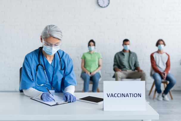mature nurse in medical mask writing on clipboard near vaccination sign plate and blurred patients - Foto, Imagen