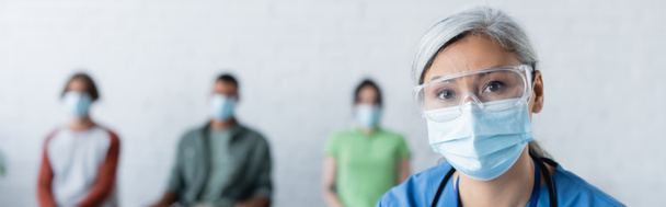 asiático médico en máscara médica y gafas mirando cámara cerca borrosa pacientes, concepto de vacunación, bandera - Foto, imagen