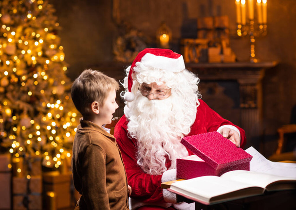 Santa Claus and little boy. Cheerful Santa is working while sitting at the table. Fireplace and Christmas Tree in the background. Traditional Christmas concept. - Foto, Imagem