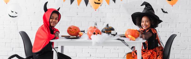 happy african american kids in halloween costumes sitting at table with pumpkins and decoration, banner  - Photo, Image