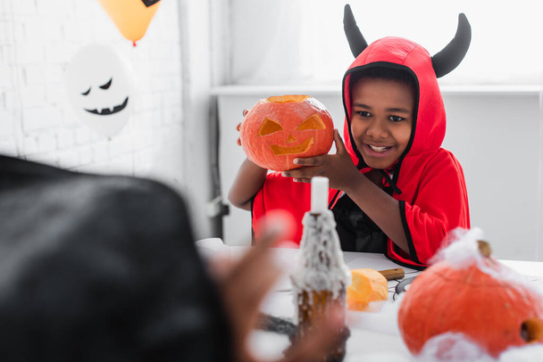happy african american boy in halloween costume holding carved pumpkin near blurred sister - Фото, изображение