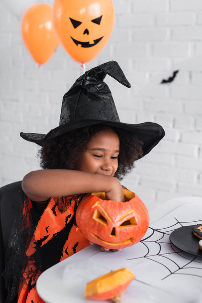 cheerful african american girl in witch halloween costume holding caved pumpkin   - Foto, afbeelding