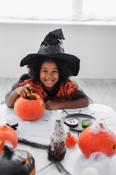 happy african american girl in witch halloween costume carving pumpkin near cookies and candles  - Foto, Imagem