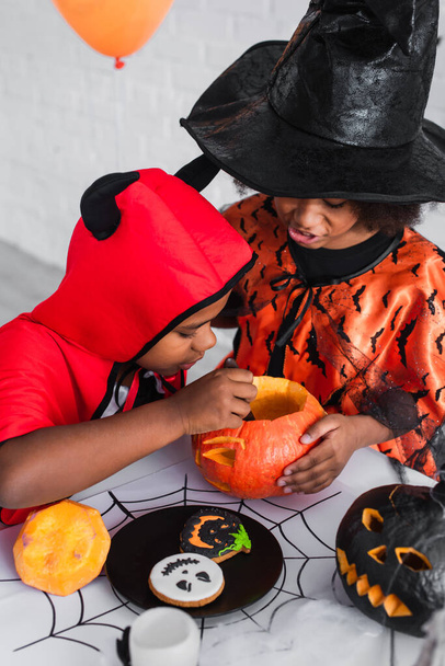 Afrikaans-Amerikaanse kinderen in halloween kostuums snijden pompoen in de buurt van koekjes  - Foto, afbeelding