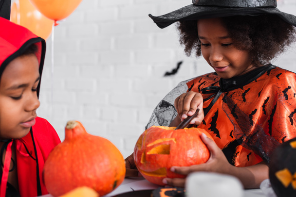 happy african american girl in pointed hat carving pumpkin near brother  - Foto, afbeelding