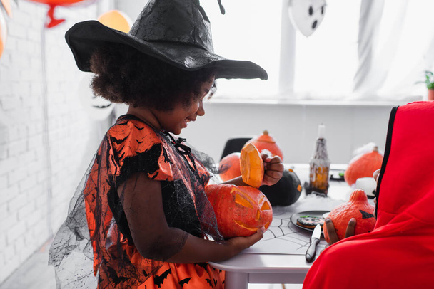 happy african american girl in pointed hat holding carved pumpkin near brother  - Photo, Image
