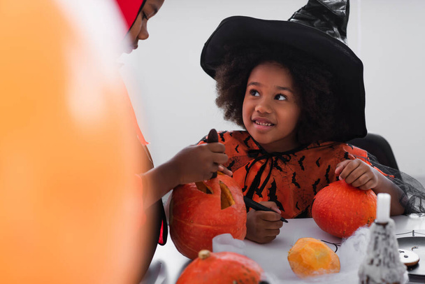 heureux afro-américaine fille en costume de sorcière regardant frère sculpter citrouille  - Photo, image