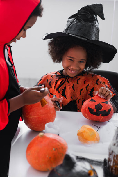 happy african american girl in witch costume showing drawing on pumpkin near blurred brother  - Zdjęcie, obraz
