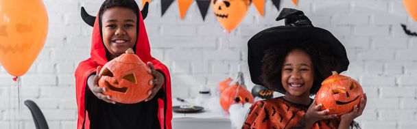 cheerful african american kids in halloween costumes holding carved pumpkins at home, banner - Φωτογραφία, εικόνα