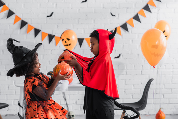 vista lateral de crianças americanas africanas em trajes de Halloween segurando abóboras esculpidas em casa - Foto, Imagem