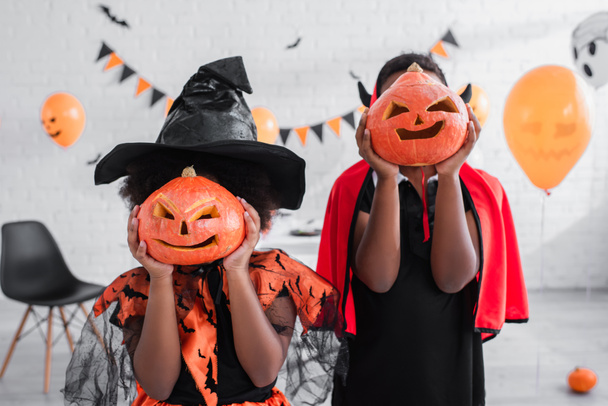 african american kids in halloween costumes covering faces with carved pumpkins at home - Фото, зображення