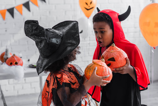 spooky african american kids in halloween costumes holding carved pumpkins at home - Zdjęcie, obraz