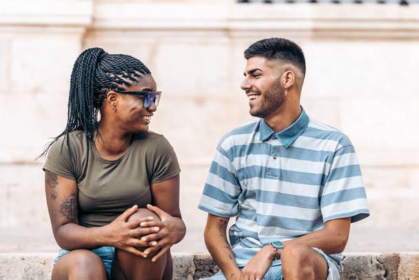 Black woman with tattoos and sunglasses sitting next to a man laughing outdoors - Foto, afbeelding