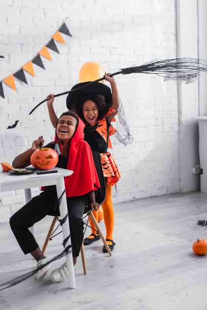 happy african american boy in halloween costume carving pumpkin near cheerful sister with broom  - Photo, Image