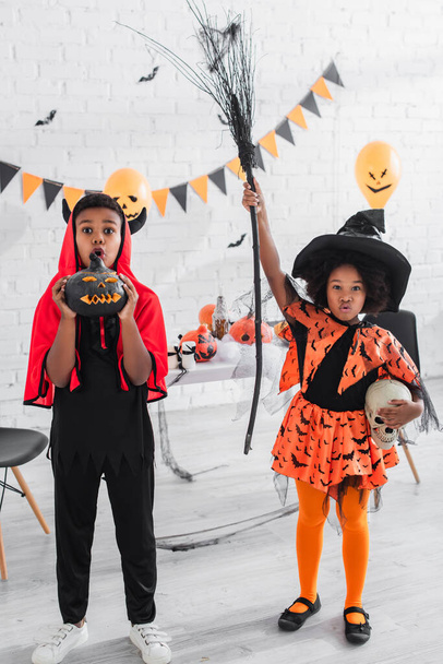 Gruseliges afrikanisch-amerikanisches Mädchen im Halloween-Kostüm mit Totenkopf und Besen neben Bruder mit geschnitztem Kürbis  - Foto, Bild