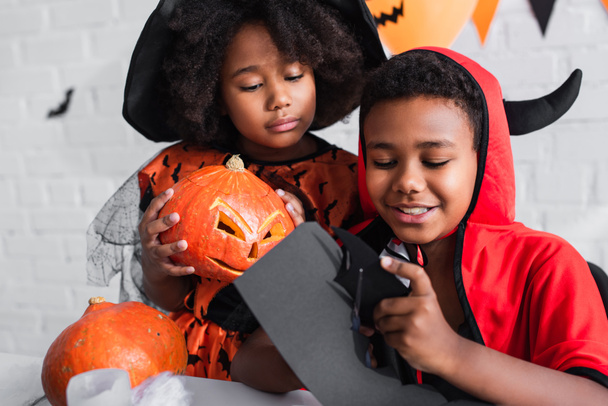счастливый африканский американец в костюме Хэллоуина режет коробку ножницами рядом с сестрой с резной тыквой  - Фото, изображение
