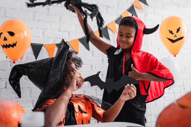 Afrikaans amerikaans meisje in halloween kostuum houden papier gesneden vleermuis in de buurt gelukkig broer met speelgoed spin tonen duim omhoog  - Foto, afbeelding