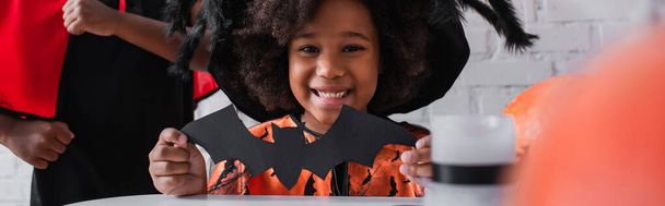 fröhliches afrikanisch-amerikanisches Mädchen im Halloween-Kostüm mit Papierfledermaus in der Nähe von Bruder, Banner - Foto, Bild