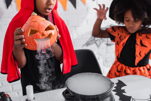 african american boy in halloween costume holding carved pumpkin with smoke while preparing potion near blurred sister  - Photo, Image