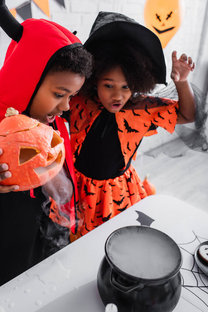 Afrikanisch-amerikanischer Junge im Halloween-Kostüm mit geschnitztem Kürbis und Rauch beim Zubereiten von Trank in der Nähe seiner Schwester mit Zipfelmütze  - Foto, Bild