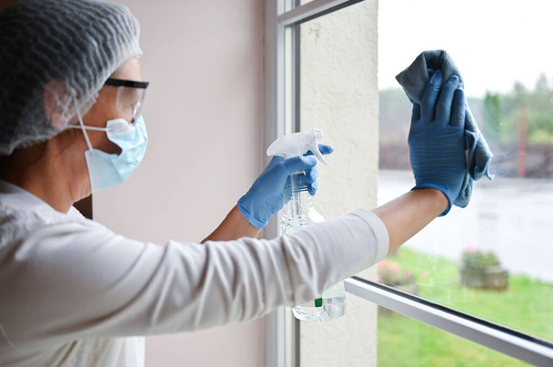 Mulher madura em terno médico protetor e máscara respiradora desinfetando móveis e vidros de janela de limpeza no hospital - Foto, Imagem