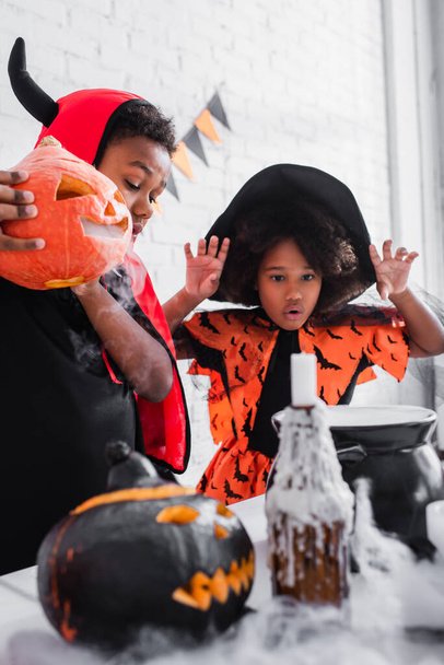 Afrikaans amerikaans jongen in halloween kostuum houden gesneden pompoen terwijl de voorbereiding van toverdrank in de buurt spooky zus in puntige hoed  - Foto, afbeelding