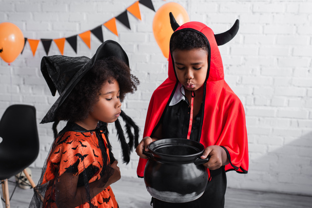 africký americký chlapec v halloween kostým držení čarodějnice kotel s lektvarem v blízkosti sestra ve špičatém klobouku - Fotografie, Obrázek