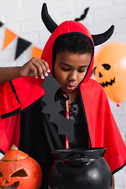 african american boy in devil halloween costume holding paper cut bat above cauldron - 写真・画像