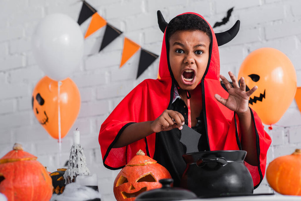 spooky african american boy in devil halloween costume holding paper cut bat above cauldron - Photo, Image