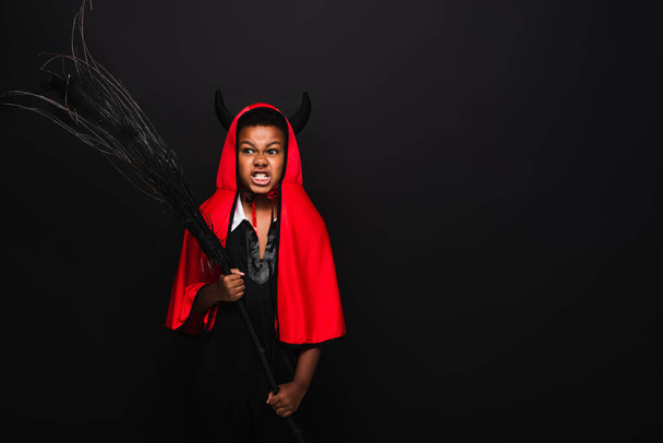 irritato africano americano ragazzo tenendo scopa e sorridendo isolato su nero  - Foto, immagini