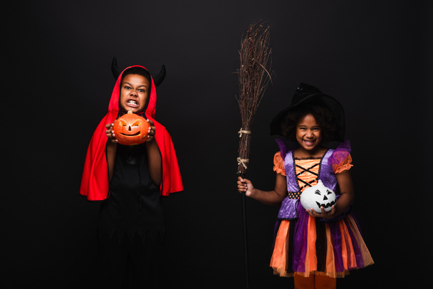 niños afroamericanos en disfraces de halloween sosteniendo calabazas talladas aisladas en negro  - Foto, imagen