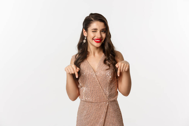 Elegant female model with red lips and earrings, pointing fingers down at christmas offer, winking and smiling, showing promo, standing in party dress over white background - Photo, image