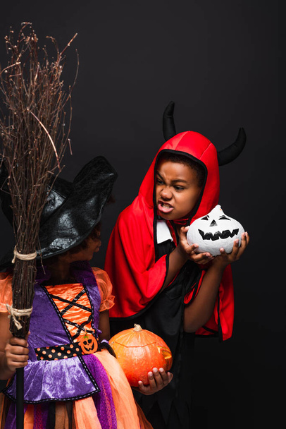 niños afroamericanos en disfraces de Halloween sosteniendo calabazas espeluznantes aislados en negro  - Foto, imagen