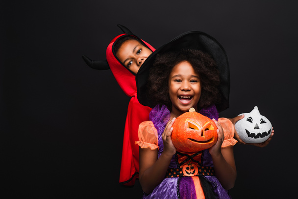 cheerful african american kids in halloween costumes holding spooky pumpkins isolated on black  - 写真・画像