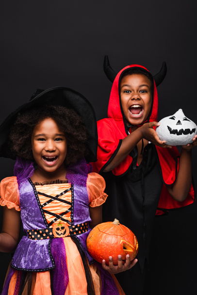 vrolijk afrikaanse amerikaanse kinderen in halloween kostuums houden griezelige pompoenen geïsoleerd op zwart  - Foto, afbeelding