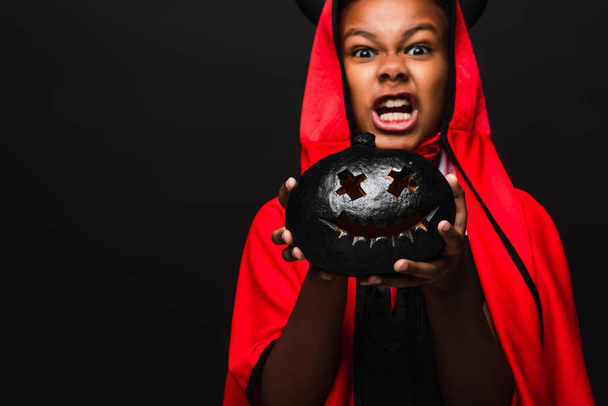 spooky african american boy in devil costume holding dark carved pumpkin isolated on black - Photo, Image