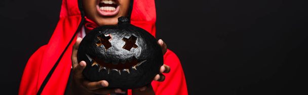 cropped view of spooky african american boy in costume holding dark carved pumpkin isolated on black, banner - Foto, afbeelding