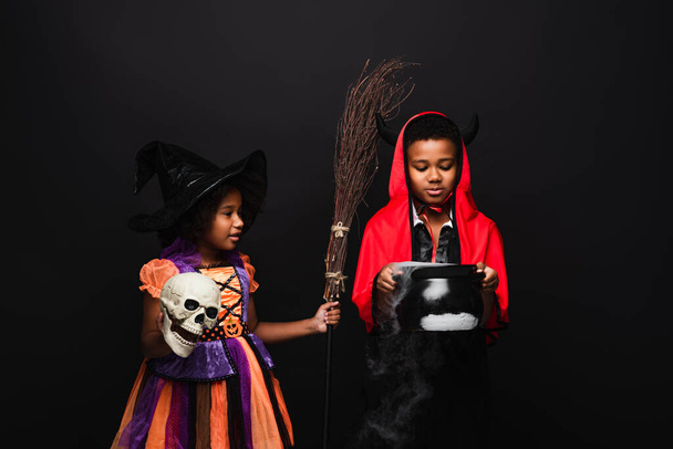 Afrikaans-Amerikaanse kinderen in halloween kostuums houden schedel en heks ketel met toverdrank geïsoleerd op zwart  - Foto, afbeelding